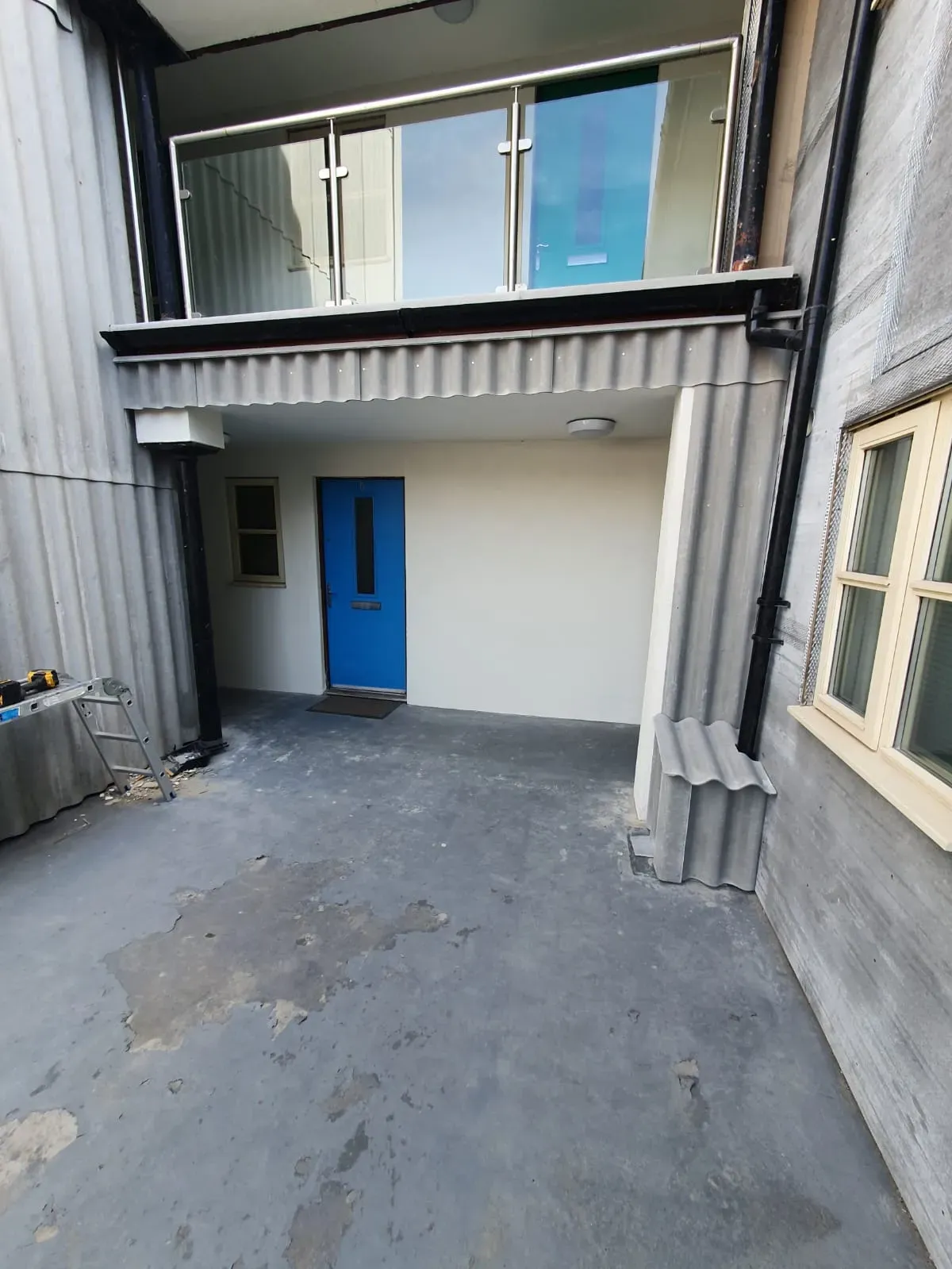 Modern building exterior showing a blue door, corrugated roofline, black downpipe, and a glass balcony.