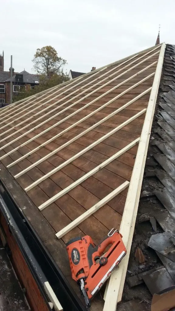 A pitched roof with new wooden battens laid across the underlay, part of a new roof installation or re-roofing project, with old roofing removed on one side.