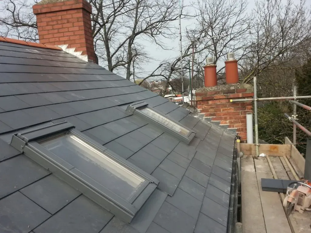 A modern dark grey tiled pitched roof with two newly installed Velux-style roof windows. In the background, brick chimneys and scaffolding are visible, suggesting recent or ongoing roofing work.