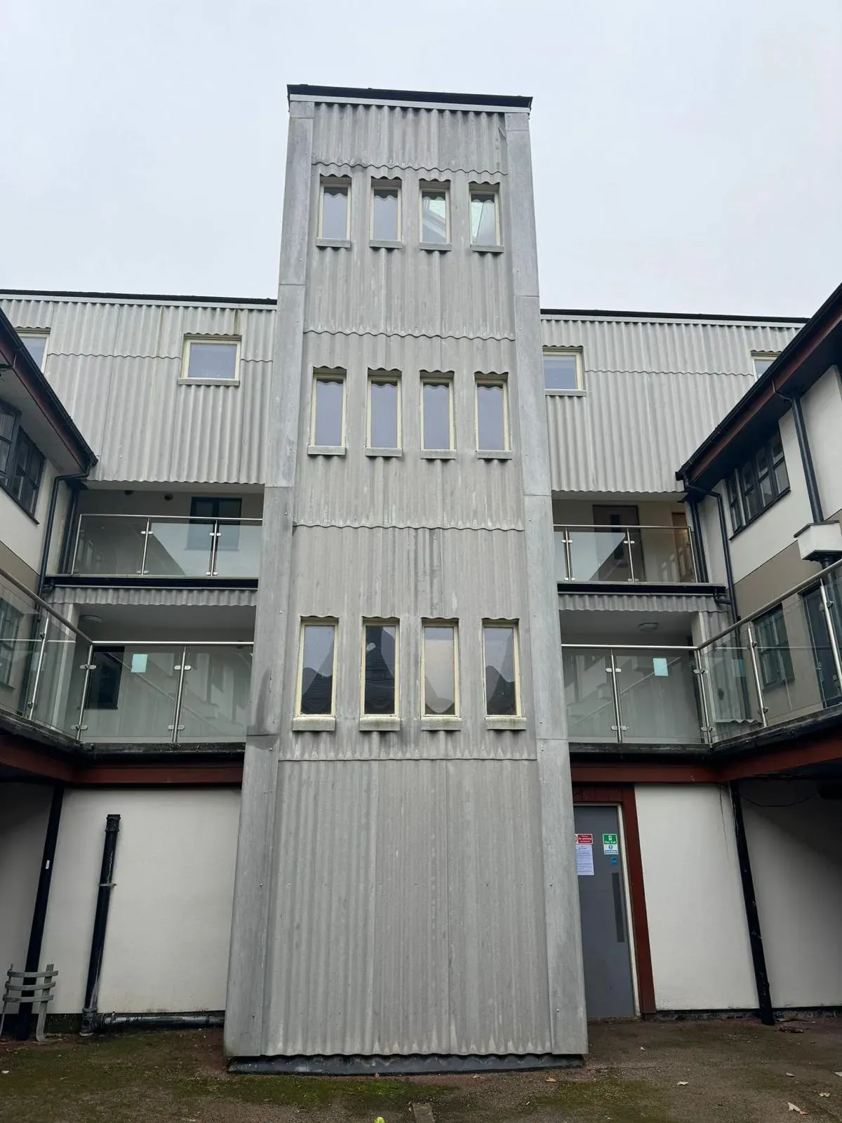 A modern building with grey corrugated fibreglass cladding and a flat roof, featuring balconies with glass railings.
